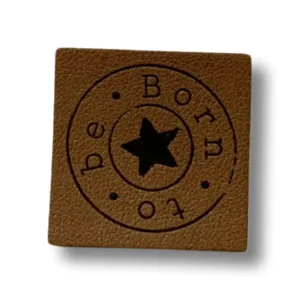 quadratisches Lederlabel mit einem schwarzen, kreisförmigen Aufdruck mit Schriftzug "born to be". In der Mitte ein schwarzer Kreis umrandet.